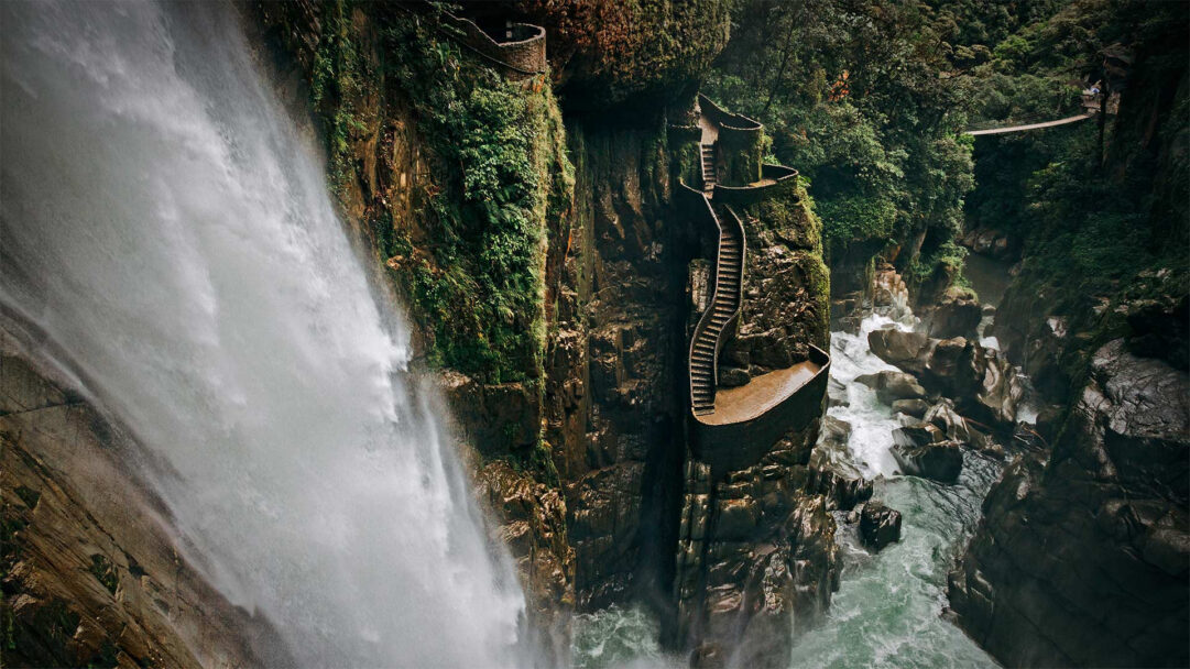 A majestic 4K wallpaper displaying the Agoyán Waterfall cascading powerfully down moss-covered cliffs in Baños de Agua Santa, Ecuador, adjacent to a winding stone staircase built into the rock face. The dramatic interplay of the forceful white water, the dark, verdant rock, and the turbulent river below evokes a sense of wild natural beauty and adventurous exploration.
