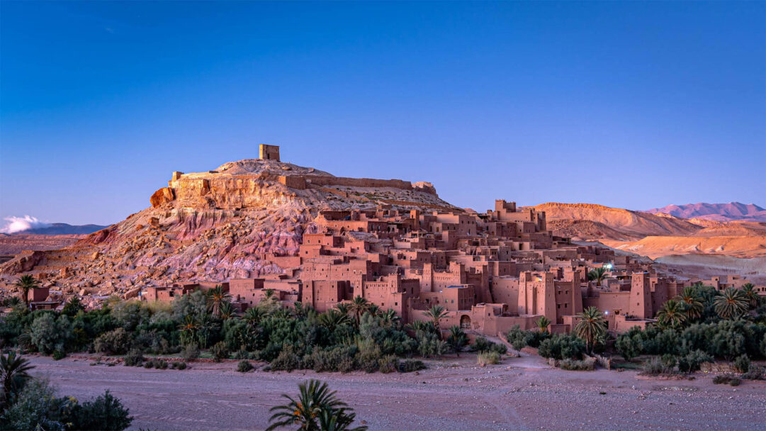 An awe-inspiring 4K wallpaper capturing the ancient Aït Benhaddou Fortified Ksar Village, nestled within the arid landscape of the Atlas Mountains in Morocco. The warm, golden light of dawn or dusk bathes the earthen buildings and rugged hillside in hues of terracotta and rose, creating a majestic and timeless atmosphere.