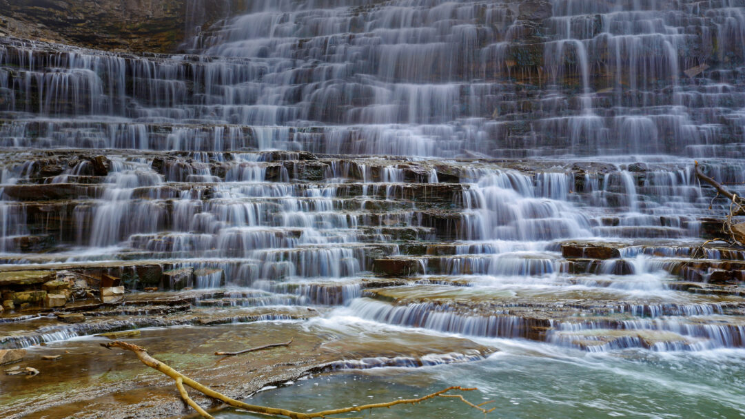 An ethereal 4K wallpaper capturing Albion Falls, a magnificent tiered waterfall in Hamilton, Ontario, Canada, with water gracefully descending over numerous rock ledges. The long exposure photography transforms the cascading water into silky white streams, beautifully contrasting with the dark, rugged rock formations and evoking a tranquil, timeless atmosphere.