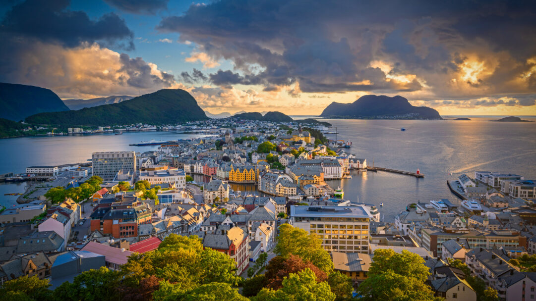 A captivating 4K wallpaper presenting Ålesund's distinctive Art Nouveau cityscape nestled amongst islands and fjords in Norway. The dramatic sunset bathes the scene in warm golden hues, highlighting the colorful buildings and reflecting vividly off the calm harbor waters, creating a serene and picturesque mood.