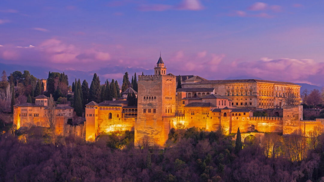 A breathtaking 4K wallpaper of the historic Alhambra Palace in Granada, Spain, majestically perched on a hillside at twilight. Its ancient walls glow brilliantly with warm golden illumination against a dramatic gradient sky of deep purples and soft pinks, creating a serene and timeless atmosphere.