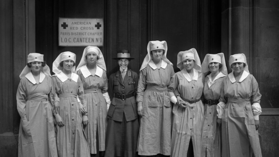 A historical 4K wallpaper showcasing a group of American Red Cross nurses and an officer proudly standing outside the Paris District Chapter L.O.C. Canteen N°1. Their vintage uniforms, crisp white caps adorned with red crosses, and the central officer's warm smile evoke a sense of dedicated service and camaraderie in this striking black and white portrait.