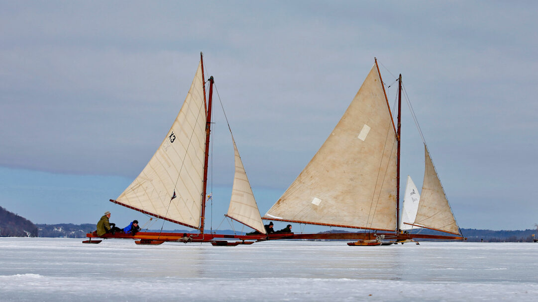 An historical 4K wallpaper featuring antique iceboats with occupants gracefully moving across the expansive frozen Hudson River near Astor Point. The muted sky above accentuates the warm, aged canvas sails and the intricate patterns of the icy river surface, evoking a tranquil yet adventurous winter scene.