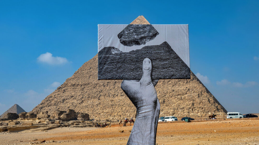 A monumental 4K wallpaper showcasing Artist JR's Pyramid Installation in the Giza desert, where a large-scale black and white cutout hand appears to hold a photographic fragment of a pyramid in front of the actual Great Pyramid of Giza. The vibrant blue sky and warm desert tones contrast sharply with the monochromatic art, creating a thought-provoking blend of ancient wonder and contemporary artistic expression.
