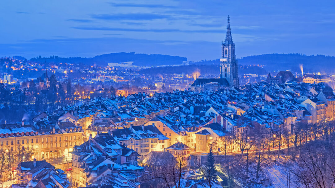 A picturesque 4K wallpaper captures the Old City of Bern, Switzerland, blanketed in fresh snow on a serene winter night. Warm golden lights emanating from windows and streetlamps illuminate the snow-dusted medieval rooftops and the soaring Bern Minster spire, creating a tranquil yet vibrant glow against the deep blue twilight.