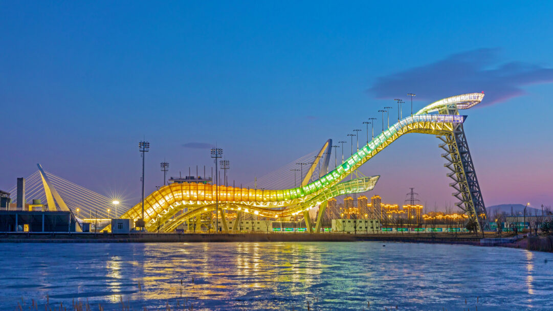 An electrifying 4K wallpaper showcasing the Big Air Shougang Olympic venue illuminated at night in Beijing, China, towering over a body of icy water. Its iconic structure glows with vibrant yellow and green lights, casting dynamic reflections across the still surface under a deep twilight sky.