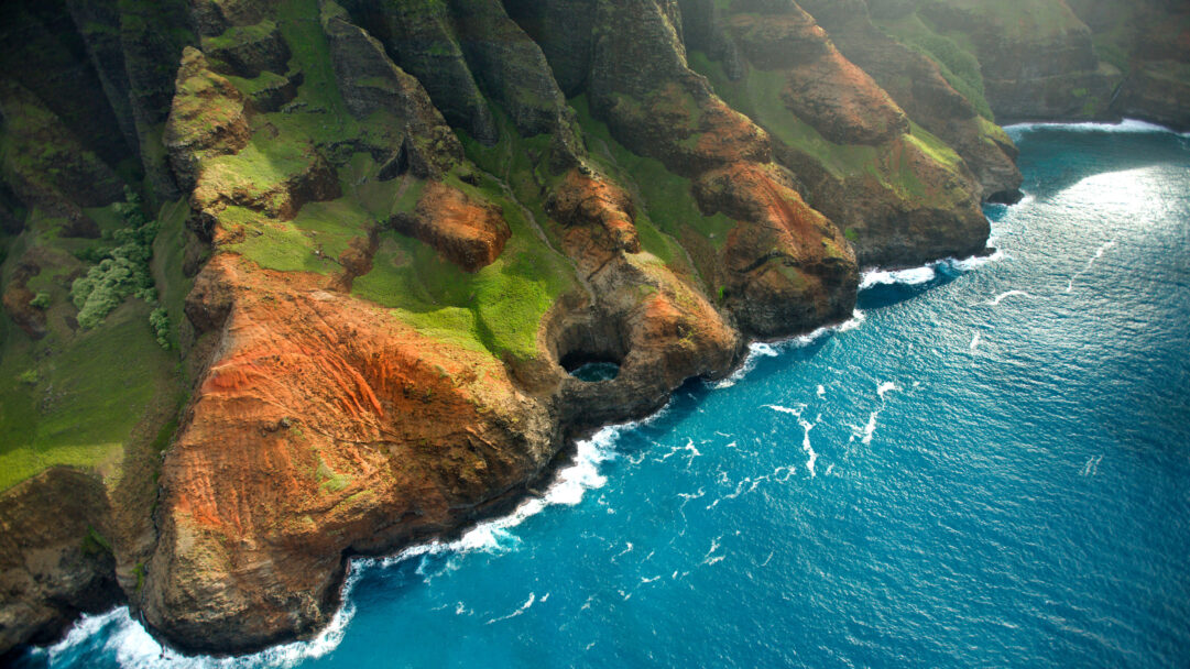 An immersive 4K wallpaper showcases the iconic Bright Eye Sea Cave nestled within the dramatic, emerald-clad cliffs of the Nā Pali Coast in Kauai, Hawaii. The intense turquoise waters of the Pacific Ocean churn around the cliff base, with sunlight illuminating the cave's deep, shadowy opening like a mysterious gaze.