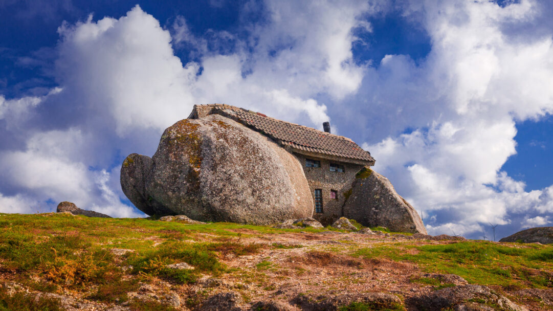 An iconic 4K wallpaper showcases the unique Casa do Penedo Stone House nestled between massive boulders on a rugged Portuguese hillside. The striking architecture blends seamlessly with the ancient, moss-covered stones, dramatically contrasting against the bright blue sky filled with billowy white clouds.