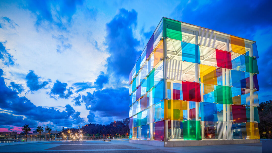 A vibrant 4K wallpaper showcasing the Centre Pompidou Málaga, a distinctive colorful glass cube architecture, situated in Spain under a dramatic evening sky. Its facade is composed of large, translucent panels in reds, blues, greens, and yellows, brightly glowing against the deep twilight clouds and distant city lights.