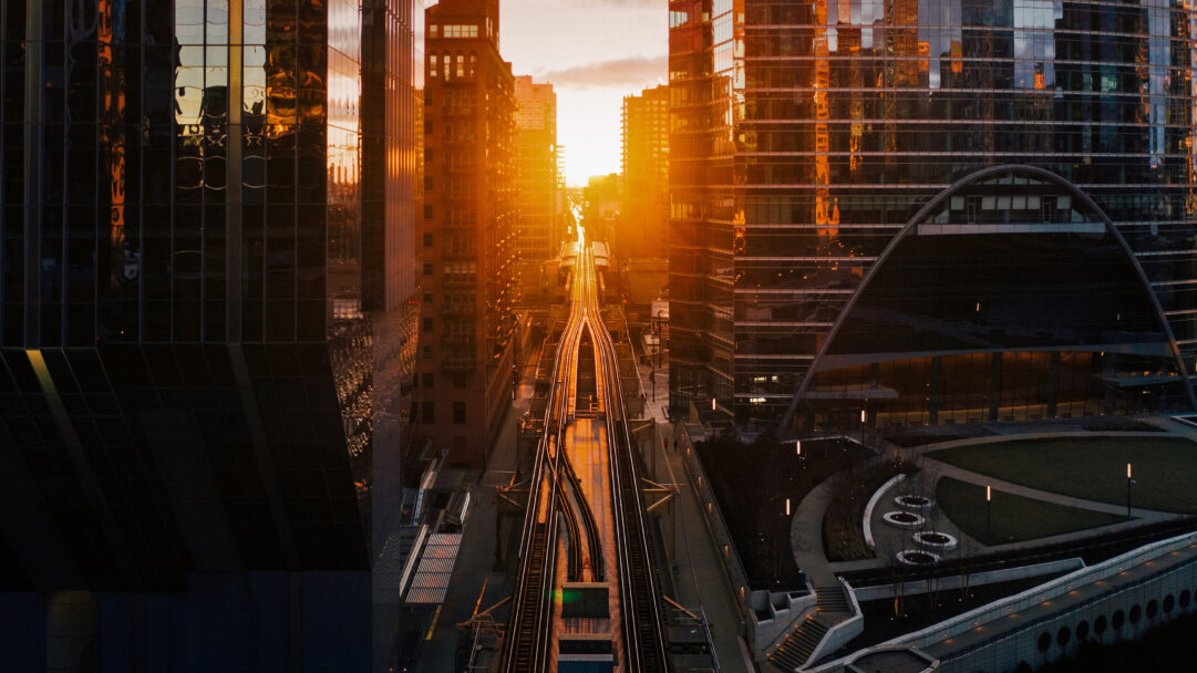 A captivating 4K wallpaper showcases the Chicagohenge phenomenon, with elevated train tracks cutting through a vibrant Chicago, Illinois cityscape. The golden sun brilliantly bathes the urban canyon and reflects off the towering glass buildings, creating a warm, dramatic glow across the city.