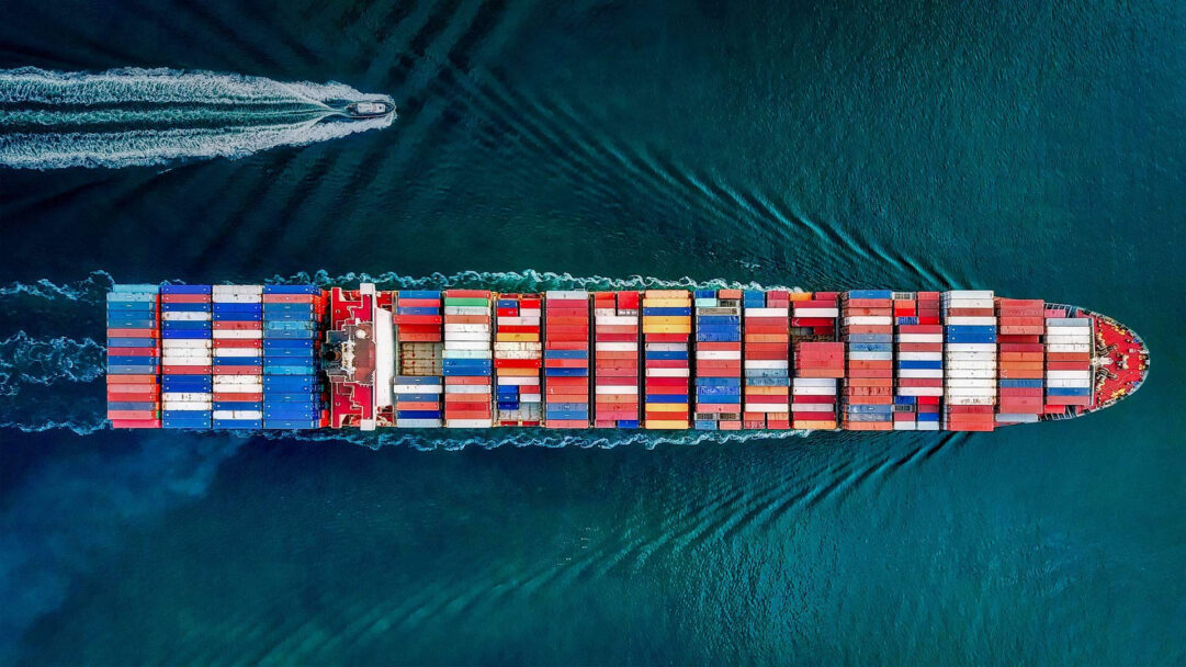A captivating 4K wallpaper presents an aerial view of a massive container ship, loaded with vibrant cargo, navigating the deep blue waters off San Pedro, California. The ship's neatly stacked, multi-colored containers create a striking mosaic against the tranquil, dark teal ocean, accented by the white foamy wakes of both the ship and a smaller escort vessel.