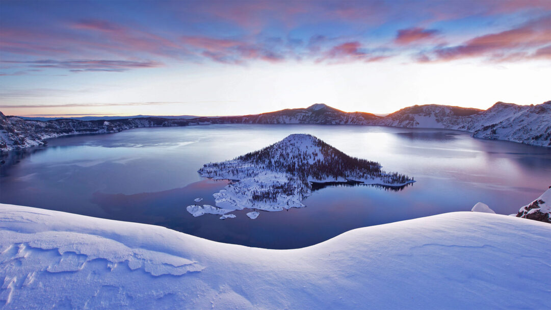 A majestic 4K wallpaper of Crater Lake, Oregon, showcasing its iconic caldera during a serene winter sunset with snow-dusted Wizard Island centered in the deep blue water. The horizon glows with soft orange and pink light, reflecting on the tranquil lake and casting a beautiful contrast against the pristine white snow covering the surrounding peaks and foreground.
