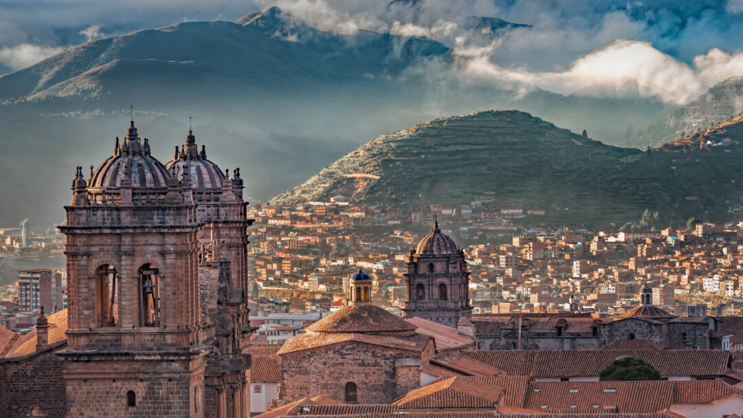 A captivating 4K wallpaper unfolds a panoramic cityscape of Cusco, Peru, dominated by the majestic Cusco Cathedral near Plaza de Armas in the foreground and stretching towards the towering Andes Mountains. Golden hour light bathes the historic terracotta rooftops of the city and the intricate stone facade of the cathedral, contrasting with the dramatic, mist-wreathed peaks of the Andes.