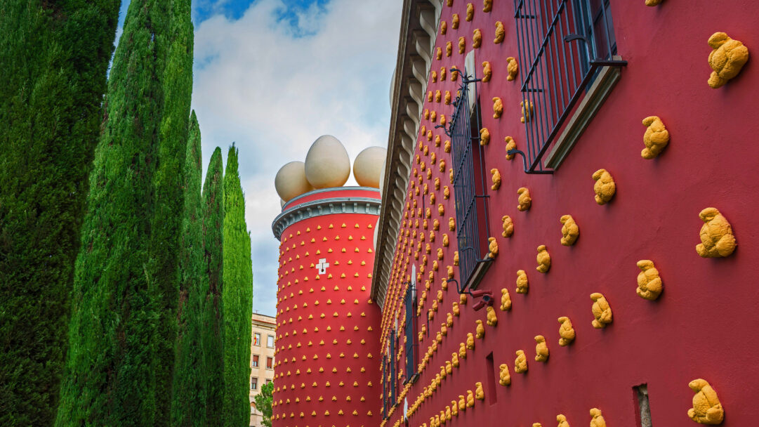 A surreal 4K wallpaper depicting the distinctive Dalí Theatre-Museum in Figueres, Spain. Its vibrant red walls are covered in a striking wallpaper of golden bread sculptures, while giant white eggs crown a prominent tower, all framed by tall green cypress trees against a bright blue sky.