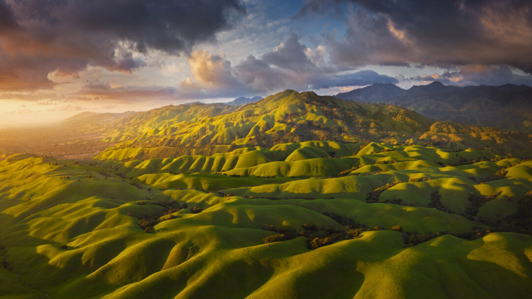 A breathtaking 4K wallpaper captures the rolling green hills of the East Bay's Diablo Range foothills, bathed in the warm glow of a vibrant sunset. Golden light illuminates the undulating contours of the hills, casting long shadows that lead up to the dramatic, colorful clouds overhead.