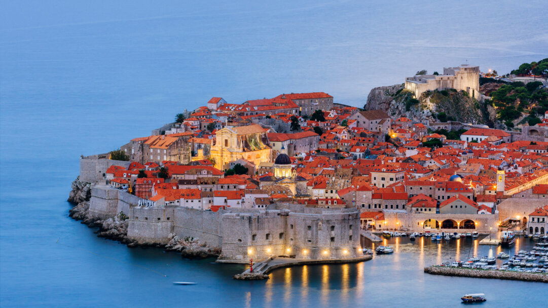A breathtaking 4K wallpaper showcases the majestic Dubrovnik Old Town in Croatia, with its ancient City Walls dramatically encircling the historic settlement as evening falls. The warm golden light from the city's myriad buildings glows against the deep blue Adriatic Sea and sky, highlighting the vibrant terracotta rooftops and creating a serene, inviting atmosphere.