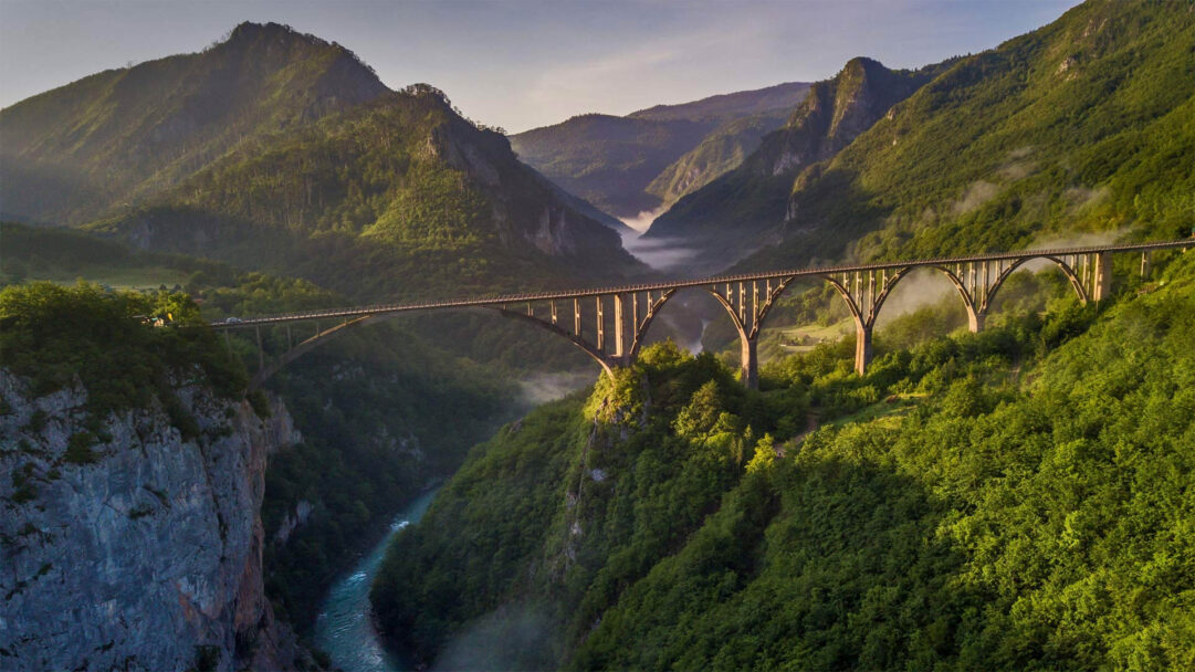 A magnificent 4K wallpaper showcasing the iconic Đurđevića Tara Bridge spanning the deep, verdant canyon of Montenegro, with a winding river flowing below. Sunlight illuminates the bridge's elegant arches and the lush forested slopes, while atmospheric mist gently fills the distant canyon, creating a serene and grand landscape.