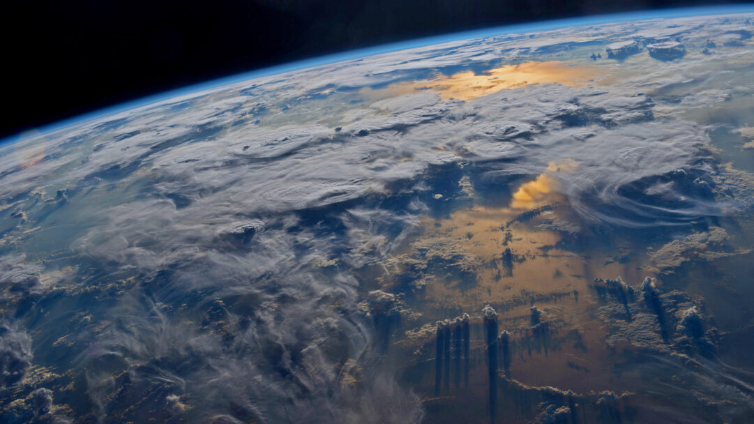 A breathtaking 4K wallpaper capturing a wide view of Earth from the International Space Station, showcasing swirling clouds over its curved horizon. Golden sunlight reflects brilliantly off dense cloud patches and a distant ocean surface, casting dramatic, elongated shadows across the planet's atmospheric layers.