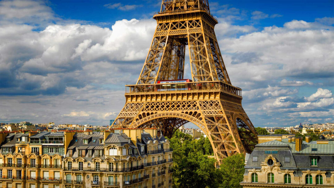 A breathtaking 4K wallpaper featuring the iconic Eiffel Tower in Paris, France, soaring above a landscape of classic city buildings. A vibrant red bus is distinctly visible within the tower's lower structure, providing a vivid contrast against the tower's intricate golden lattice and the dramatic blue sky filled with fluffy white clouds.