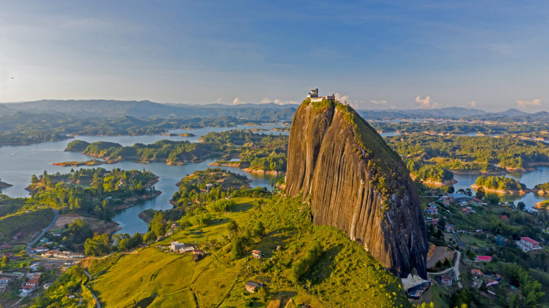 An awe-inspiring 4K wallpaper showcasing El Peñón de Guatapé, a massive granite rock monolith, rising majestically from the vibrant, island-dotted reservoir landscape of Guatapé, Antioquia. Golden hour light bathes the scene, illuminating the rock's vertical striations and the lush green hills, creating a tranquil yet grand natural vista.