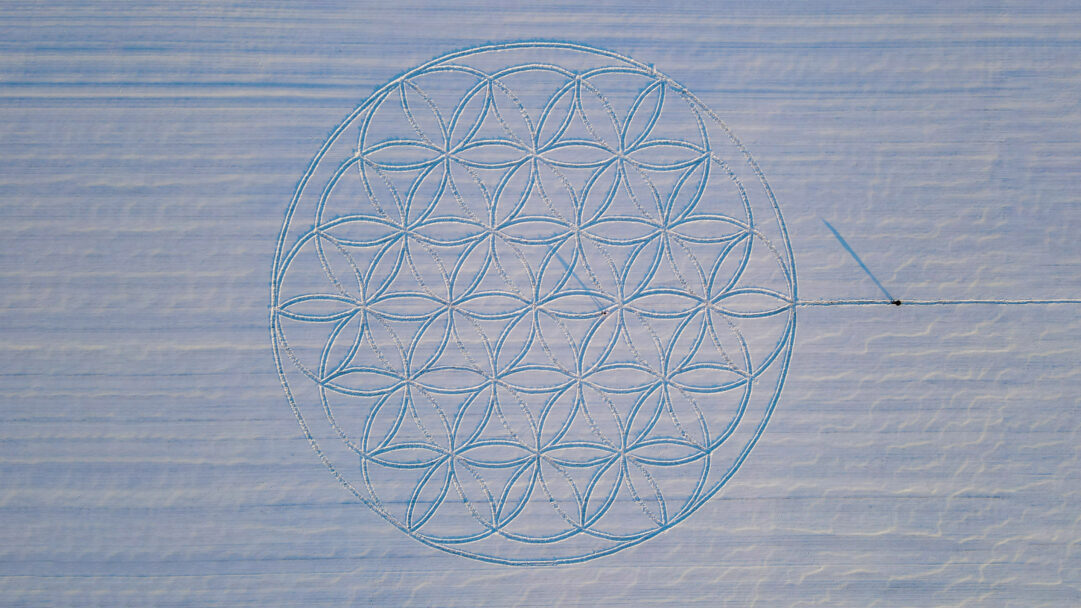 An exquisite 4K wallpaper of Michael Uy's 'Flower of Life' snow art, presented as a majestic aerial view across a pristine winter field. The vast geometric pattern is precisely etched into the snow, with a lone figure visible at its edge, creating a striking contrast of delicate artistry and expansive, tranquil scenery.
