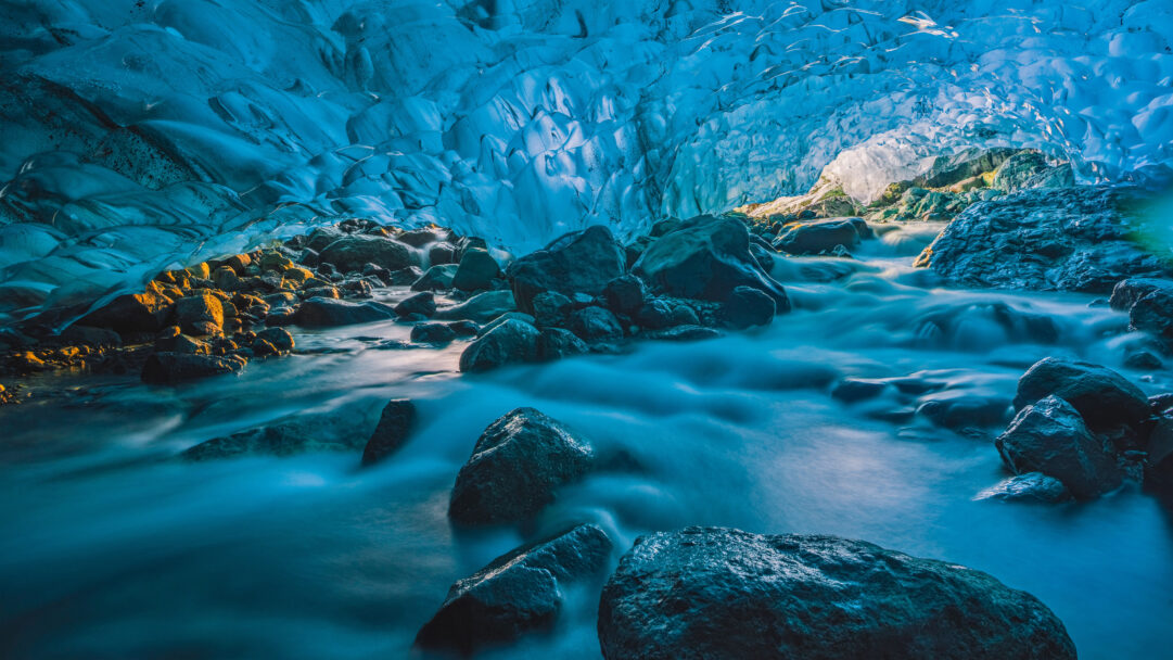 A mesmerizing 4K wallpaper reveals a flowing river winding through a majestic glacier cave in Vatnajökull, Iceland. The cave's luminous blue ice walls are beautifully illuminated by a distant golden light, contrasting with the silky, ethereal blue water flowing over dark, wet rocks.