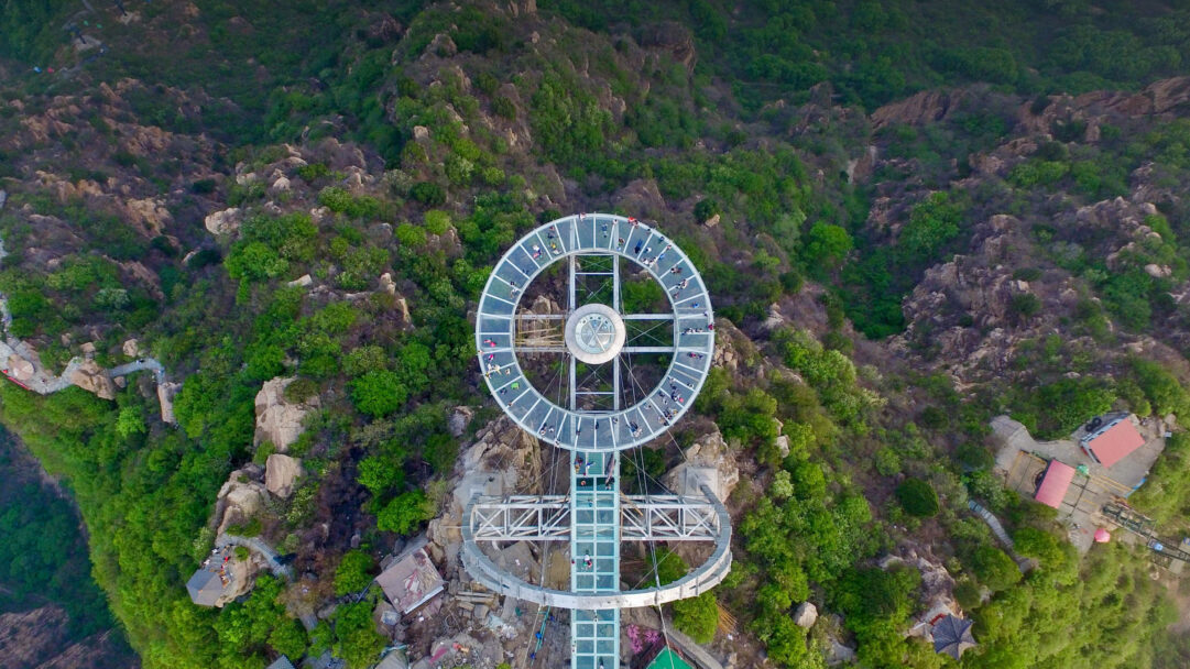 A breathtaking 4K wallpaper showcasing an aerial view of the circular Glass Sightseeing Platform at Shilinxia Scenic Area, perched high on a lush green mountain. Numerous small figures walk across the transparent glass floor, offering a dizzying perspective of the dramatic, verdant mountain terrain below.