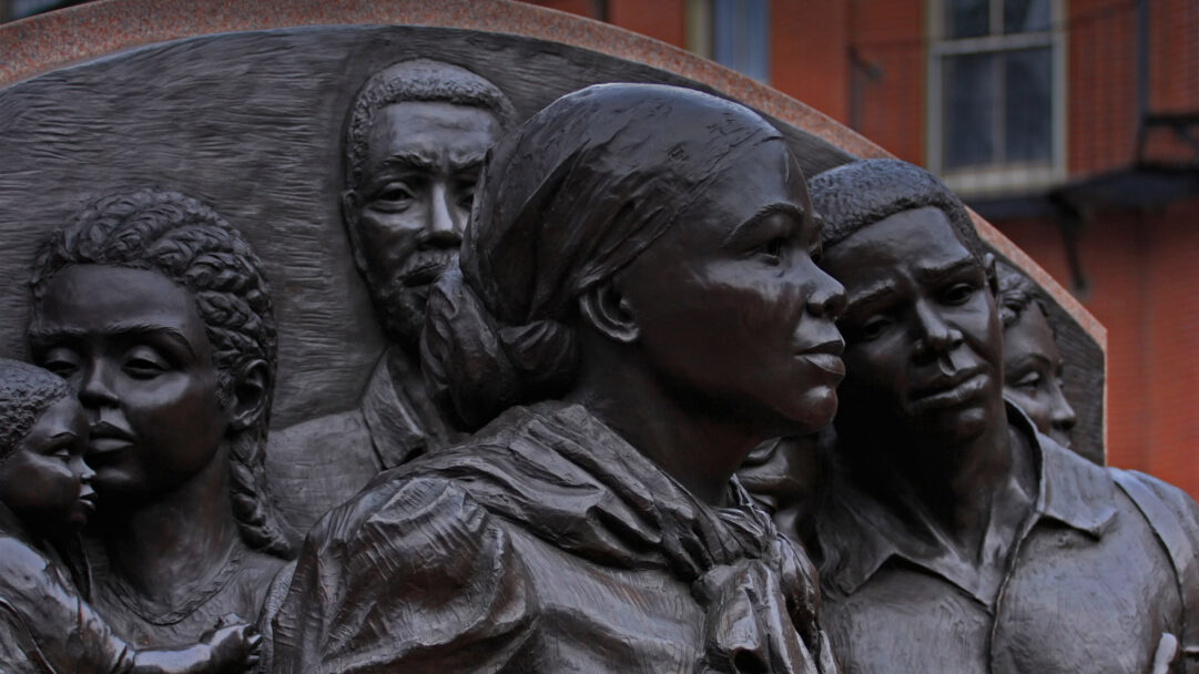 A powerful 4K wallpaper presenting the bronze Harriet Tubman Memorial Sculpture by Fern Cunningham, where Harriet Tubman leads a resolute group of figures. The deep bronze hues beautifully highlight the intricate textures and expressions of courage and determination, set against a softly blurred reddish-brown brick building.