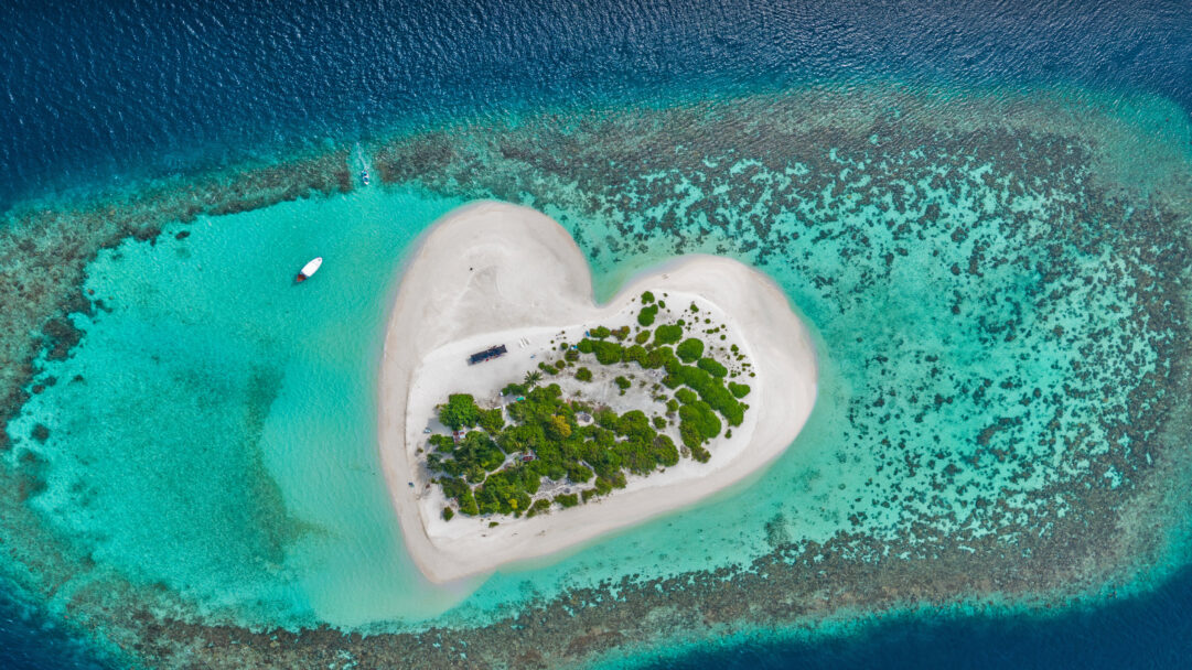 A captivating 4K wallpaper showcases a perfect heart-shaped island in the Maldives, featuring a pristine sandy beach surrounded by a vibrant coral reef within the vast Indian Ocean. The striking gradient of turquoise shallows fading into the deep blue ocean perfectly frames the lush green interior and white sands, evoking a sense of tranquil paradise.