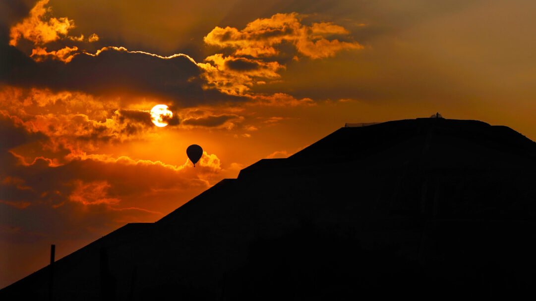 An iconic 4K wallpaper of a hot air balloon silhouetted over the Pyramid of the Sun in Teotihuacan during a dramatic sunrise. The vibrant orange and golden sky, bursting with sunlight through atmospheric clouds, creates a majestic backdrop for the dark, imposing pyramid and the lone, ascending balloon.