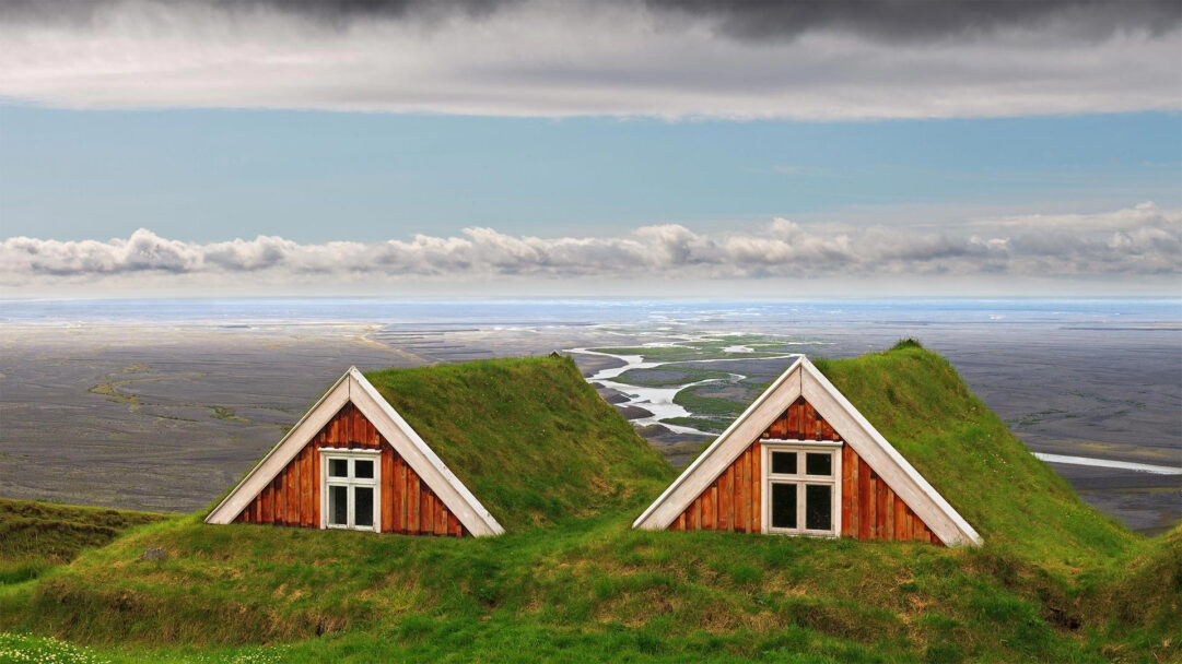 An iconic 4K wallpaper showcasing two traditional Icelandic turf houses with vibrant green roofs nestled into grassy mounds, overlooking a vast, dark river delta landscape. The striking contrast of their rustic wooden gables and lush green roofs against the expansive, braided glacial river winding through a barren, dark plain creates a serene, timeless mood.