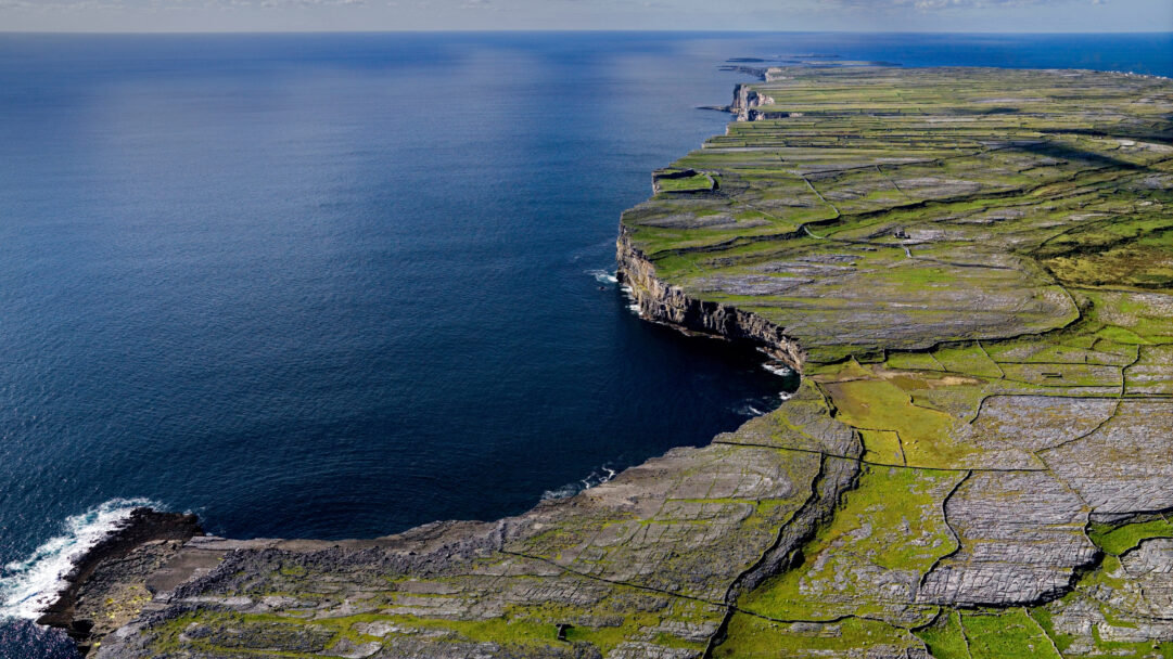A breathtaking 4K wallpaper depicts the rugged Irish coastline, where dramatic cliffs meet the vast Atlantic Ocean, extending into the distant horizon. Intricately patterned green fields, sectioned by ancient stone walls, dramatically contrast with the sheer, dark rock faces descending into the deep blue water.