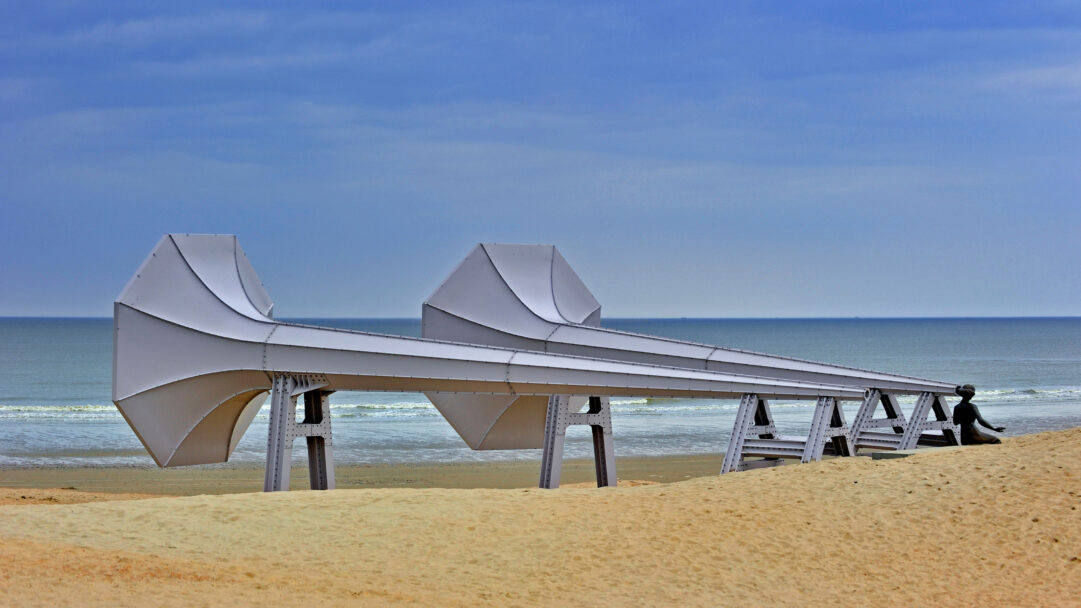 An intriguing 4K wallpaper presents Ivars Drulle's monumental gray art installation, a series of connected horn-like structures, stretching across a vast sandy beach towards the calm ocean. A solitary dark figure sits at the installation's end, quietly observing the expansive seascape under a soft blue sky.