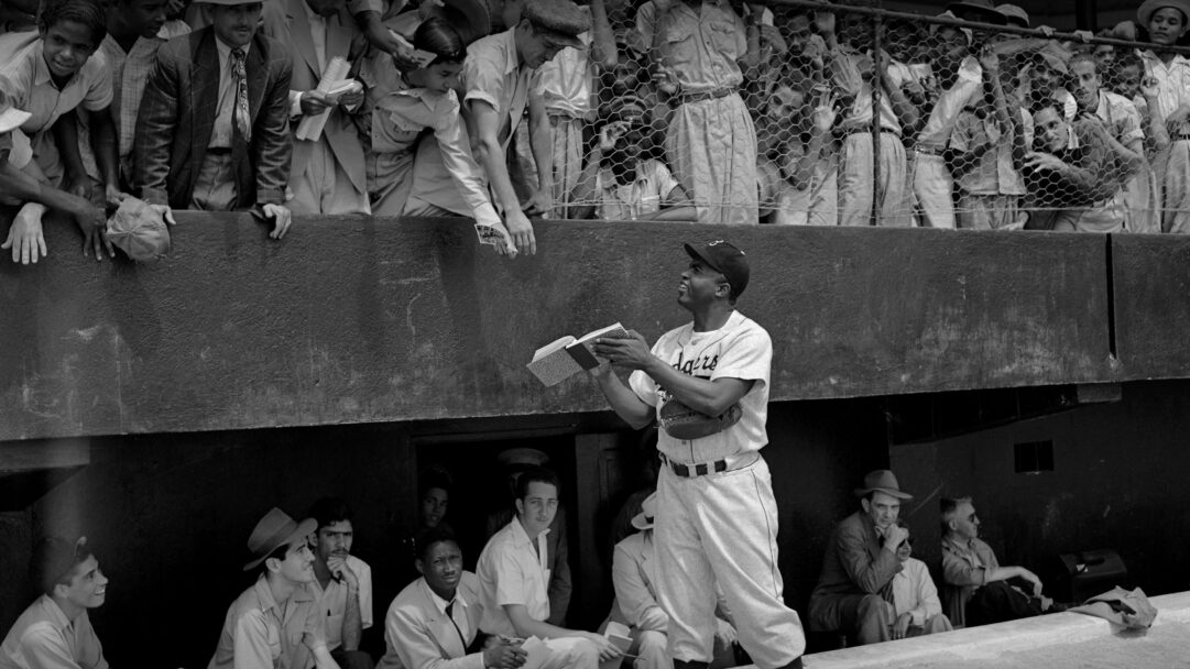 An iconic 4K wallpaper captures Jackie Robinson, in his Dodgers uniform, diligently signing autographs for enthusiastic fans at Spring Training. The black-and-white image accentuates the historical moment, showing numerous hands reaching over a barrier towards Robinson, who holds an open book, reflecting intense public admiration and the era's bustling atmosphere.