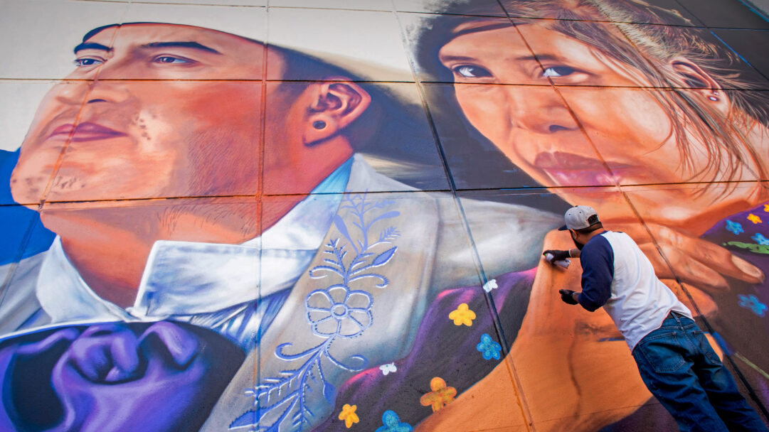 A powerful 4K wallpaper featuring artist Jesus 'Cimi' Alvarado intently painting his monumental 'Fronterizos' mural on a large wall, which depicts the expressive faces of a man and a woman. His active spray painting brings vibrant life and intricate detail to the vast artwork, emphasizing the dynamic interplay of color and texture against the wall's surface.