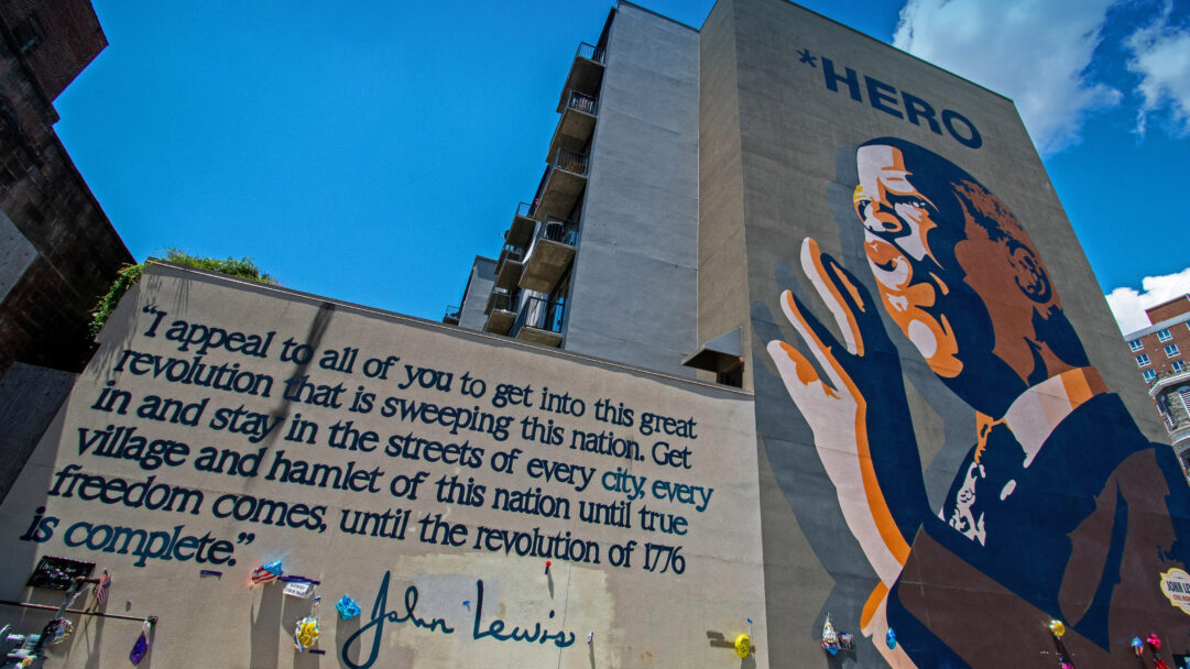 An impactful 4K wallpaper displays Sean Schwab's vibrant John Lewis Hero Mural, dominating the side of a building in Atlanta's historic Sweet Auburn District. The mural features a stylized portrait of John Lewis with raised hands, accompanied by his powerful call to action quote, inspiring a sense of continued revolution under a brilliant blue sky.