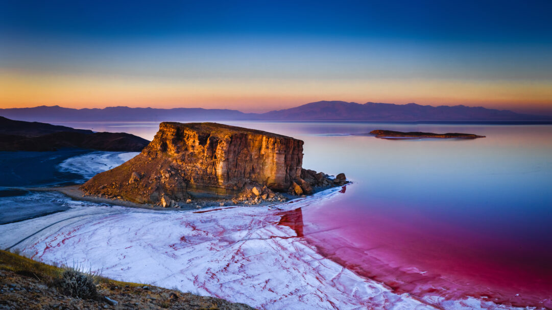 A breathtaking 4K wallpaper captures the majestic Kazem Dashi Rock Formation standing proudly in Lake Urmia, Iran, at sunset. The lake transforms into a vivid pink salt expanse, reflecting the warm hues of the setting sun, while stark white salt crusts line the shore, creating a surreal landscape.