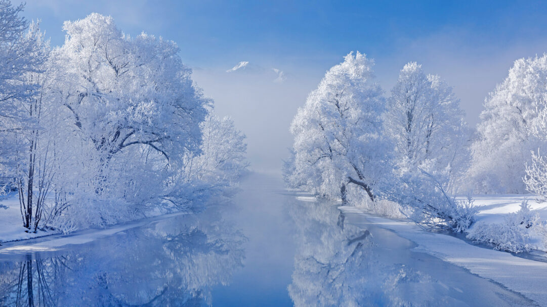 An ethereal 4K wallpaper captures Kochelsee's winter beauty in Bavaria, Germany, featuring a river flanked by trees heavy with white frost and the distant silhouette of Heimgarten Mountain. The tranquil river's reflective, icy-blue surface mirrors the snow-laden branches and clear sky, evoking a serene and pristine winter landscape.
