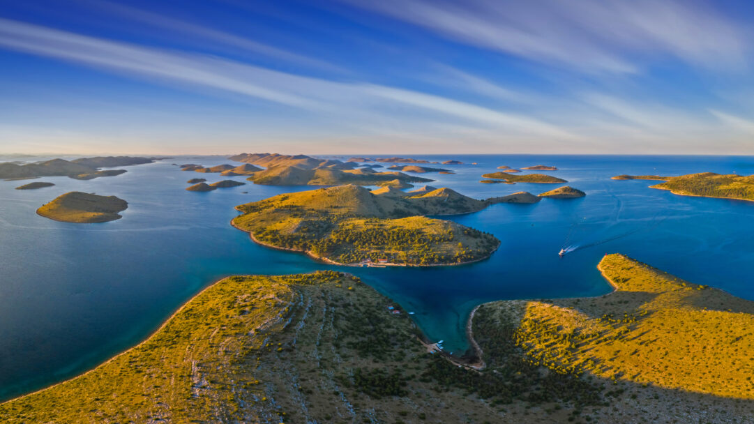 A breathtaking 4K wallpaper showcasing the sprawling Kornati National Park in Croatia, an archipelago of numerous islands spread across a deep blue seascape under a streaked sky. Golden hour sunlight dramatically highlights the arid, rocky terrain of the islands, creating striking contrasts with the brilliant turquoise and indigo waters where a boat cruises.