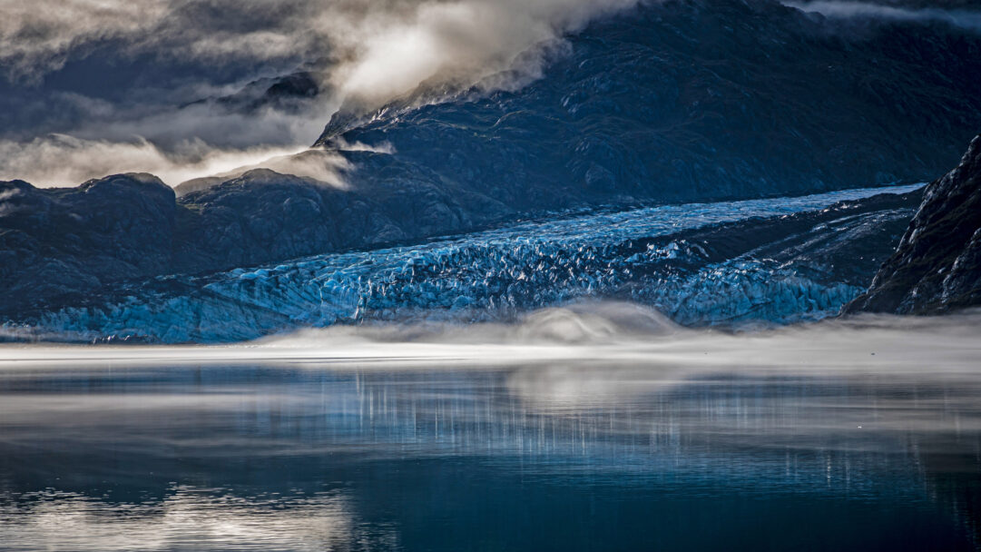 A majestic 4K wallpaper unveils Lamplugh Glacier within Alaska's Glacier Bay National Park, presenting a dramatic misty landscape where the vibrant blue glacier face meets calm, reflective waters. Atmospheric mist gently hovers above the water, contrasting with the rugged, cloud-shrouded mountains and lending an ethereal, tranquil beauty to the scene.