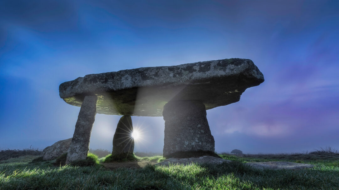 A dramatic 4K wallpaper capturing the ancient Lanyon Quoit Burial Chamber in Cornwall, England, standing on a dew-kissed grassy field beneath a striking blue sky. A brilliant sunburst emanates from behind the megalithic stones, creating a mystical interplay of light and shadow across the landscape.