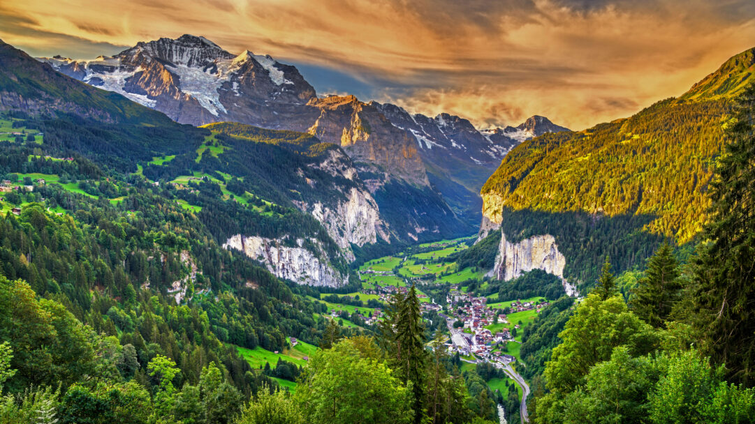 A breathtaking 4K wallpaper capturing the expansive Lauterbrunnen Valley in the Swiss Alps, where a charming mountain village is nestled amidst lush greenery. The warm glow of the sunset illuminates the dramatic snow-capped peaks and the distinctive rock faces, creating a serene and majestic mood.