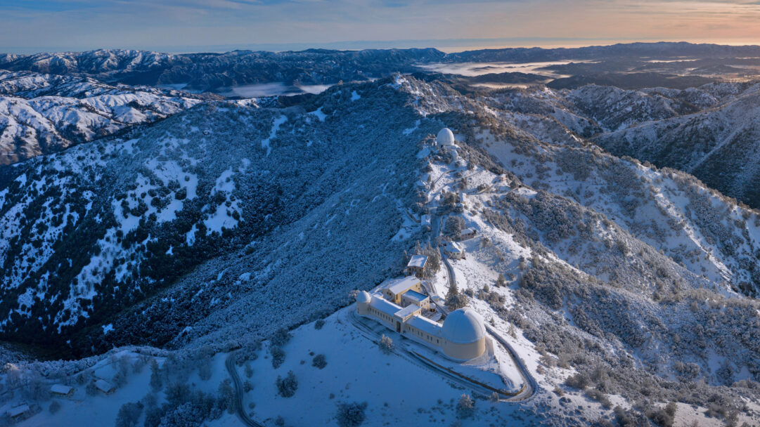 A breathtaking 4K wallpaper presenting Lick Observatory nestled atop snow-covered Mount Hamilton in San Jose, California. The crisp winter snow blankets the rugged peaks and surrounding evergreen trees, while a soft golden light graces the distant horizon over a valley filled with mist.