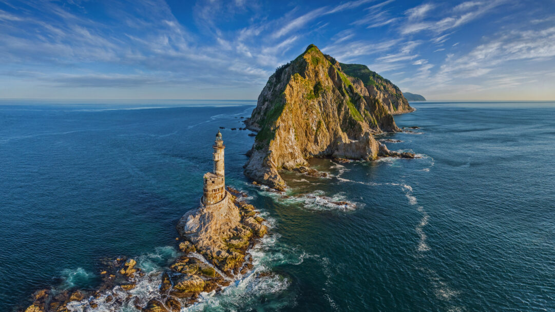A dramatic 4K wallpaper presents the iconic Lighthouse at Cape Aniva, Sakhalin Island, Russia, standing on a rugged rocky islet in the expansive ocean. Golden sunlight illuminates the lighthouse's aged, decaying structure and the colossal, green-topped cliffs behind it, creating a powerful sense of isolated majesty against the deep blue, foam-laced sea.