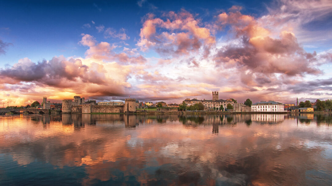 A captivating 4K wallpaper depicting Limerick, Ireland's historic cityscape stretching along the River Shannon at sunset. The vivid orange and pink clouds overhead are beautifully mirrored in the calm water, highlighting the city's ancient stone architecture with a warm, serene glow.