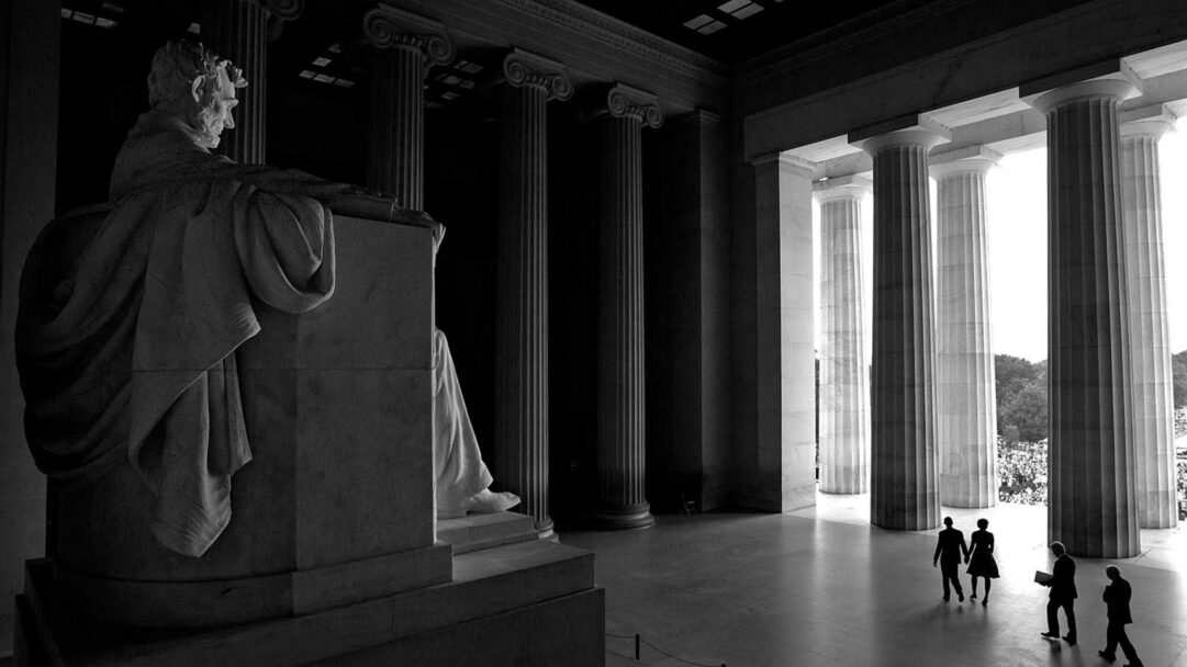 A profound 4K wallpaper featuring the colossal seated statue of Abraham Lincoln inside the grand, columned hall of the Lincoln Memorial in Washington D.C. The grayscale composition emphasizes the stark contrast between the statue's contemplative shadows and the brilliant light streaming through the massive exterior columns, drawing small silhouetted figures towards the entrance.