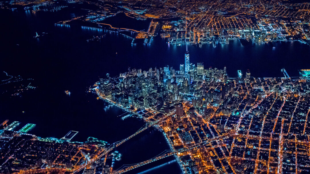A breathtaking 4K wallpaper showcases a vibrant aerial view of Lower Manhattan, New York City, extending across the dark waters at night. The urban landscape is defined by the sparkling grid of lights, with the iconic One World Trade Center standing tall amidst the warm and cool hues reflecting on the dark harbor.