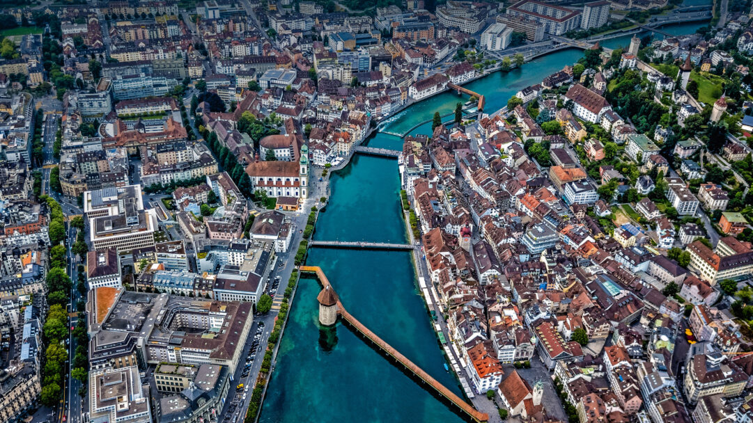 An expansive 4K wallpaper showcasing an aerial cityscape of Lucerne, Switzerland, where the vivid turquoise Reuss River carves through a dense arrangement of historic and modern buildings. The famed Chapel Bridge and its octagonal Water Tower stand prominently in the river, their warm wooden tones contrasting beautifully with the cool, deep blues of the water and the varied rooftops.