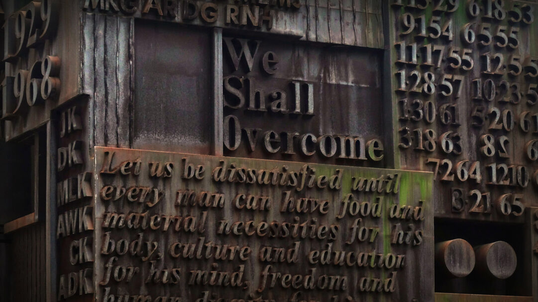 A profound 4K wallpaper showcases a close-up of William Tarr's MLK Memorial Sculpture, prominently displaying the words We Shall Overcome in raised, dark metal. The aged bronze surface, rich with texture and further etched quotes advocating for human dignity, evokes a powerful sense of historical struggle and enduring hope.