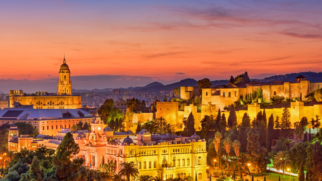 A captivating 4K wallpaper showcases Málaga Old Town's sprawling cityscape in Spain, highlighting the majestic Alcazaba and Cathedral at sunset. The ancient Alcazaba and the Cathedral glow with warm golden light against a dramatic twilight sky, painted in rich hues of orange, pink, and soft purple, creating a serene and majestic atmosphere.